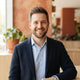 A friendly man smiling in a professional headshot with warm terracotta and white background tones.